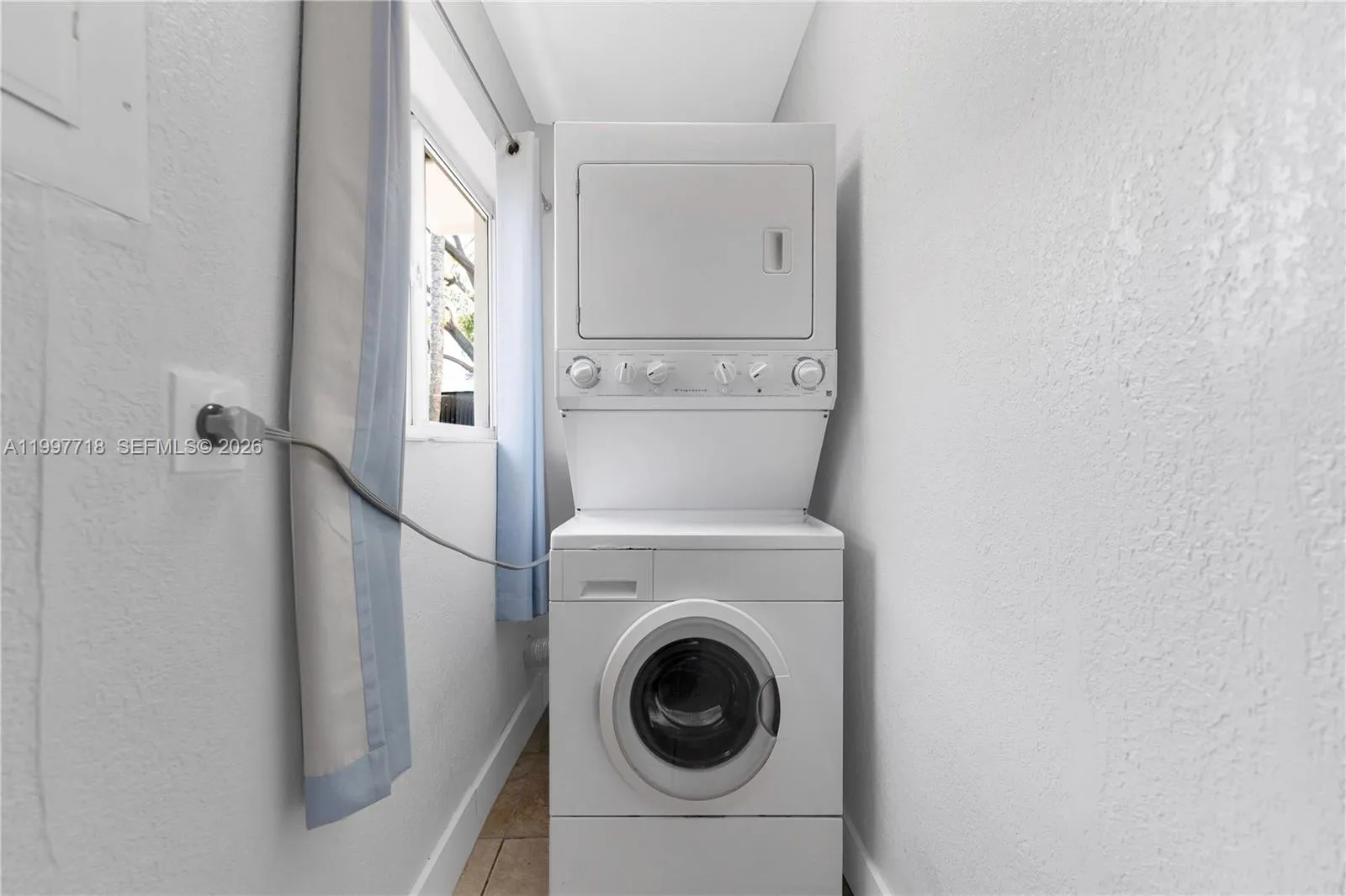 Laundry Area\r\nA narrow laundry nook with a stacked washer and dryer unit tucked against the wall, tile flooring, and a small window with a light blue curtain.