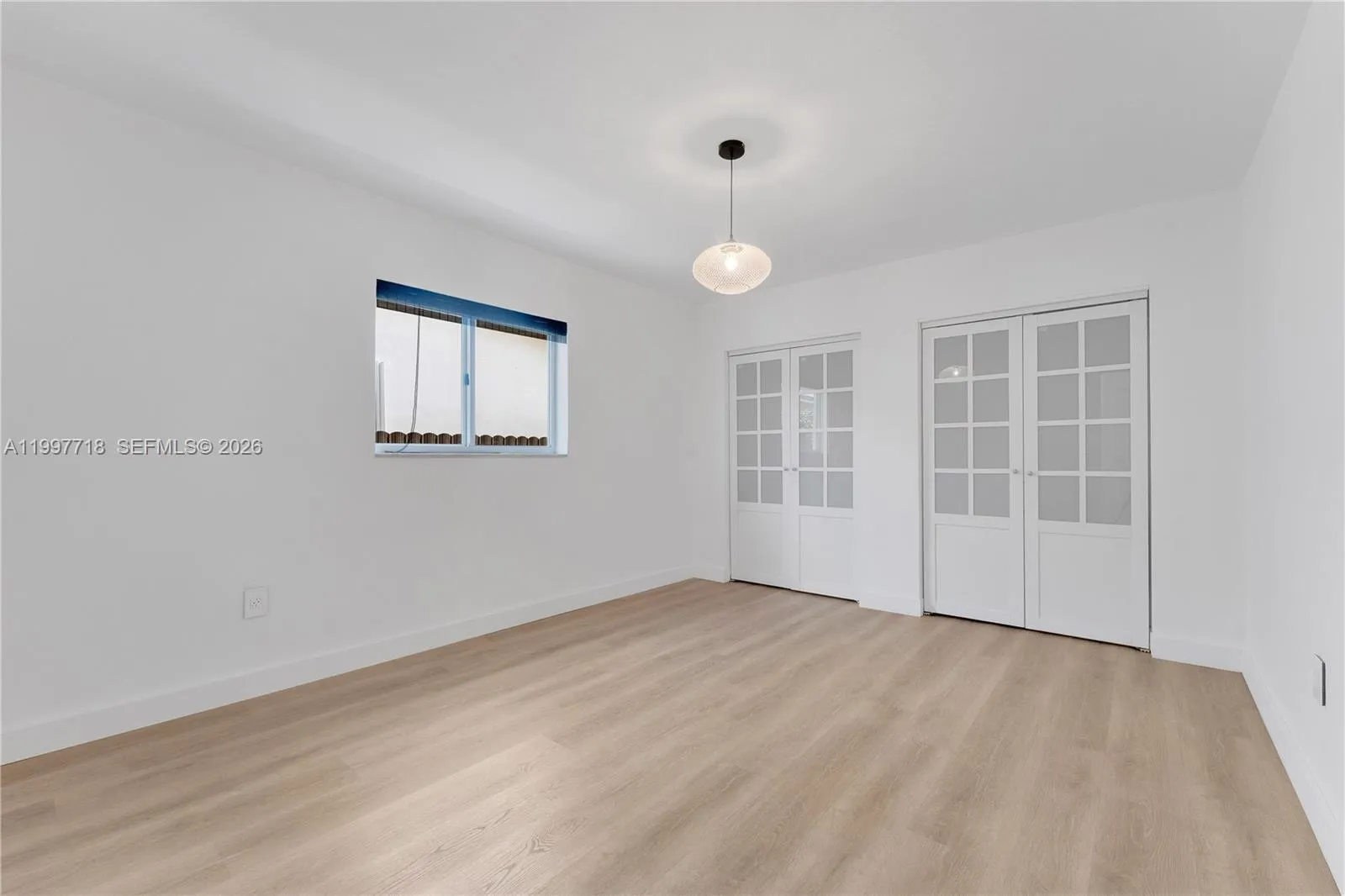 A clean, empty room with light hardwood-style flooring, white walls, a stylish pendant light, a side window with a roller shade, and double French glass doors leading to the main living area.