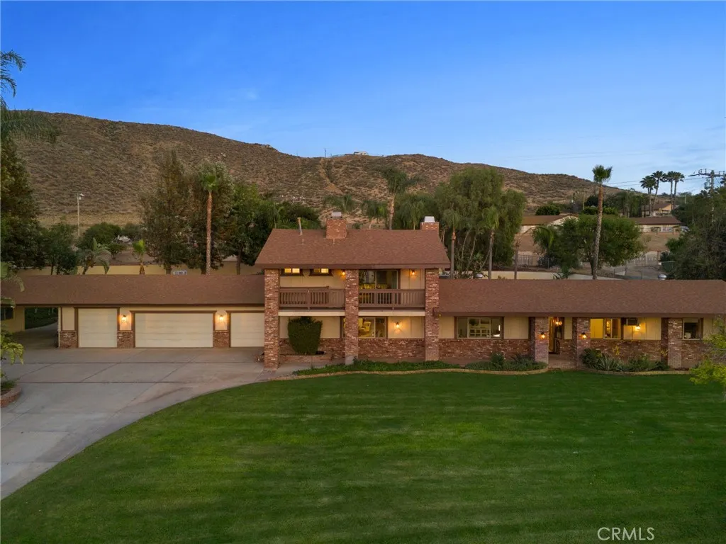 Long brick-and-stucco home with a two-story center, attached garages, and a broad lawn beneath dry hills.