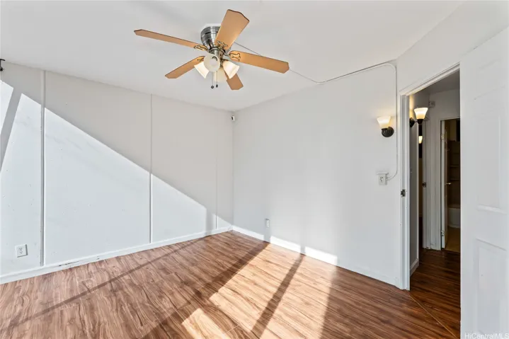 A bright bedroom with wood flooring, white walls, a ceiling fan with light, and an open doorway leading to the bathroom.