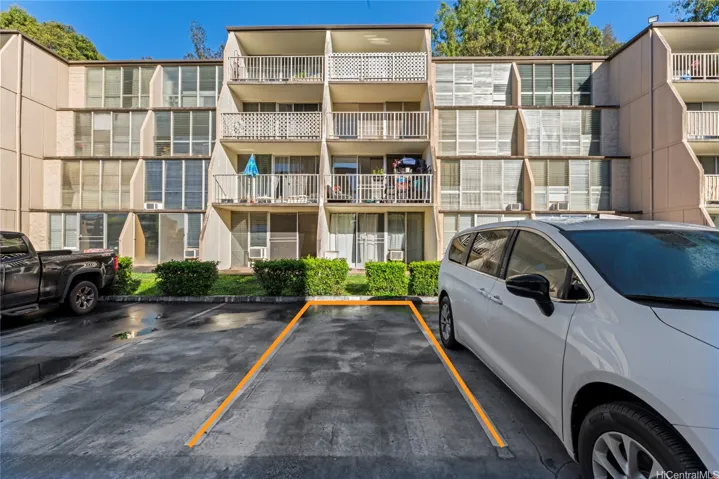 An exterior view of the residential building with balconies, assigned parking spaces, and landscaped greenery in front.