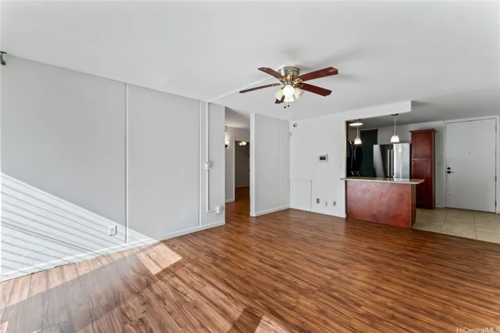 A bright, open living space featuring warm wood floors, neutral walls, a ceiling fan, and a partial view of the kitchen with a breakfast bar.