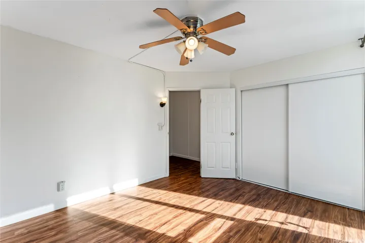 Another angle of the room showing wood flooring, a ceiling fan with light, sliding closet doors, and an open doorway.