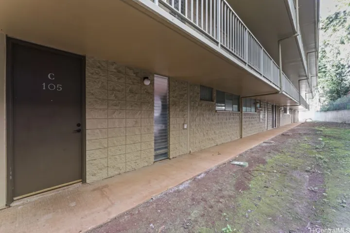 An exterior ground-level walkway of the building showing unit entrances, covered balconies above, and a landscaped area alongside the path.