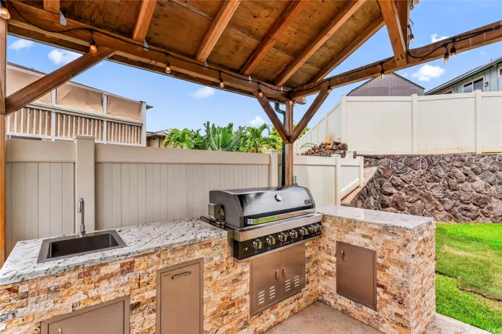Covered outdoor kitchen with a built-in grill, sink, stone counters, and string lighting.