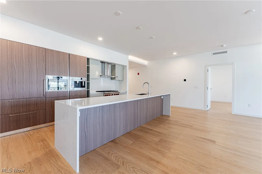 Kitchen featuring wall chimney exhaust hood, stainless steel oven, sink, light hardwood / wood-style floors, and an island with sink
