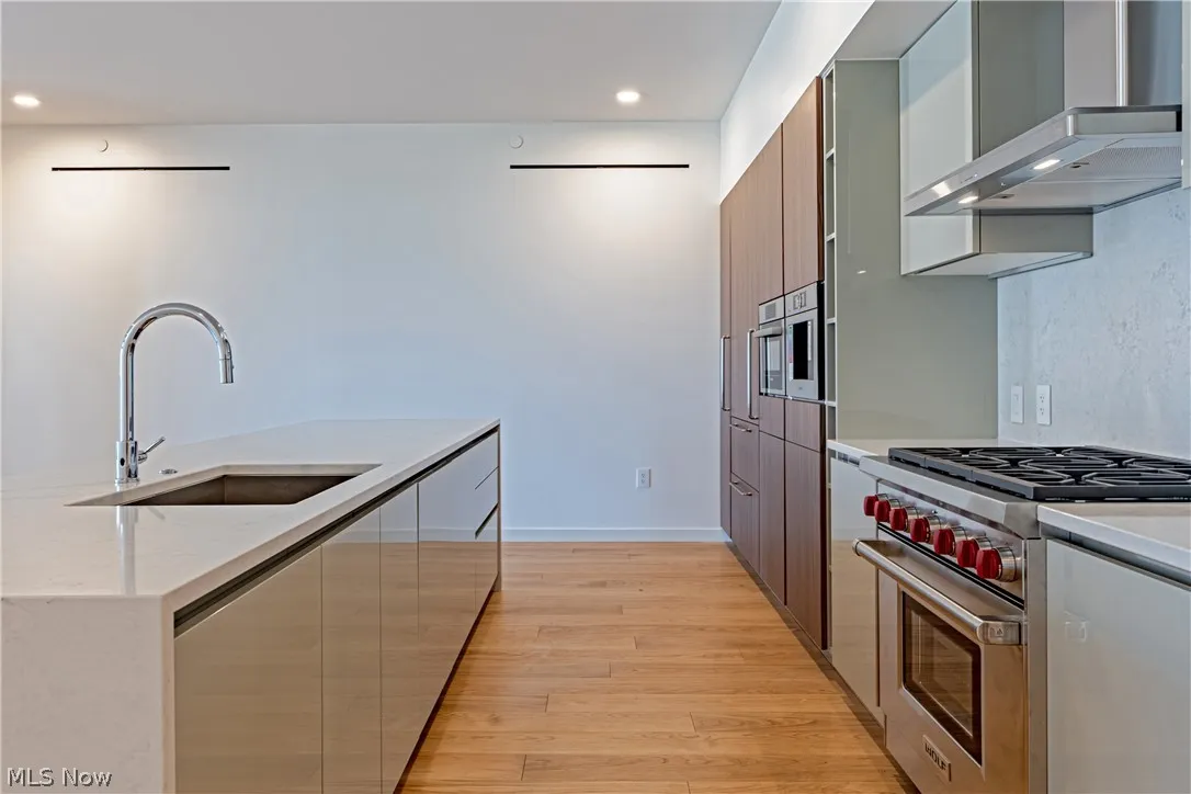 Kitchen with sink, designer range, wall chimney range hood, light wood-type flooring, and light stone countertops