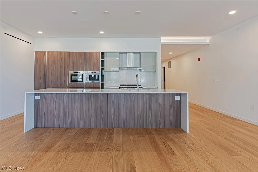 Kitchen with wall chimney range hood, light wood-type flooring, and a kitchen island with sink