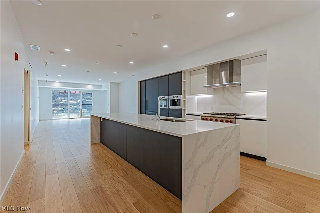 Kitchen featuring light hardwood / wood-style floors, a center island with sink, backsplash, and wall chimney exhaust hood