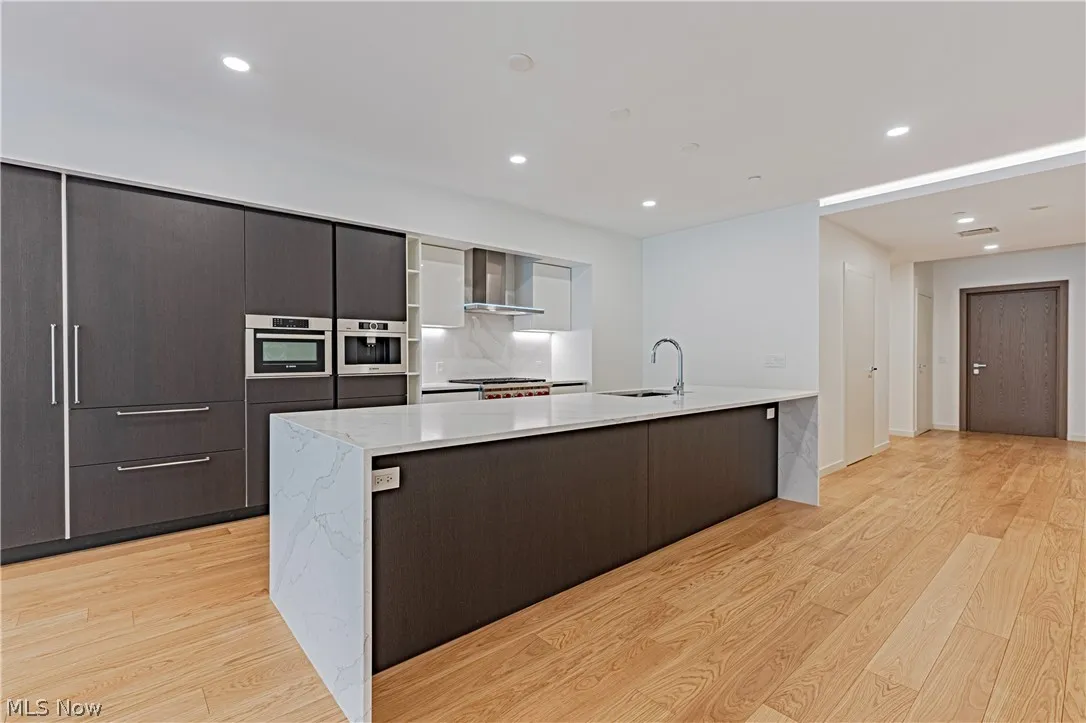 Kitchen featuring wall chimney exhaust hood, light stone countertops, light hardwood / wood-style flooring, sink, and an island with sink