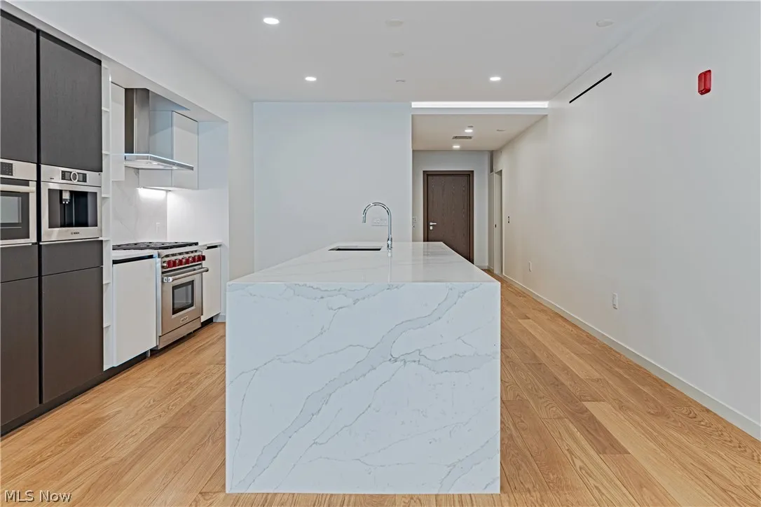 Kitchen featuring wall chimney exhaust hood, light hardwood / wood-style flooring, stainless steel appliances, sink, and a center island with sink