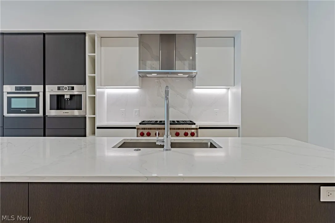 Kitchen featuring wall chimney range hood, stainless steel oven, gray cabinets, and light stone countertops