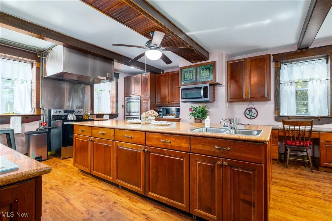 Kitchen featuring ceiling fan, light wood-type flooring, wall chimney exhaust hood, a center island with sink, and appliances with stainless steel finishes
