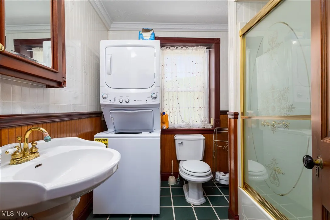 Full bathroom with tile patterned floors, stacked washing maching and dryer, ornamental molding, sink, and toilet