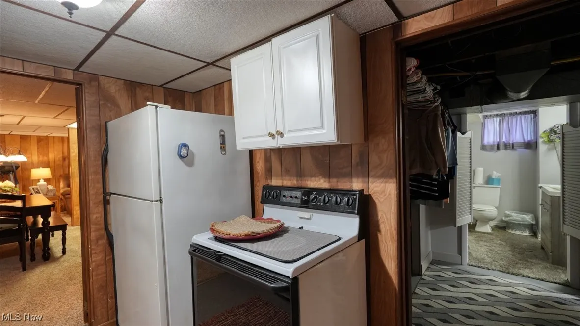 Kitchen featuring a paneled ceiling, light carpet, wood walls, white cabinetry, and white appliances