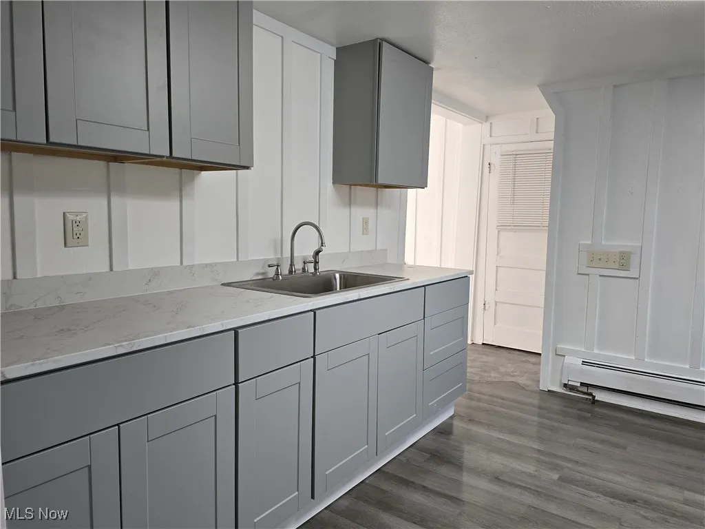 Kitchen with gray cabinets, dark hardwood / wood-style floors, a baseboard radiator, sink, and light stone countertops