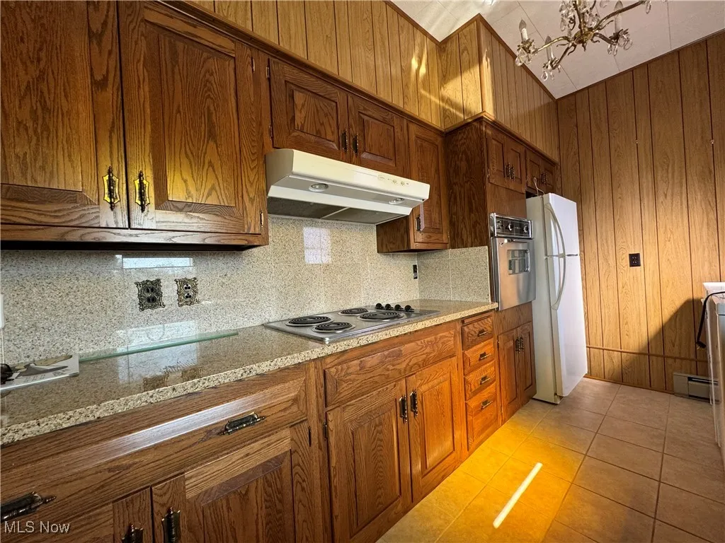 Kitchen featuring backsplash, light tile patterned floors, light stone counters, stainless steel appliances, and a notable chandelier