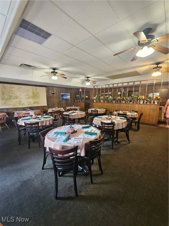 Dining room with wooden walls, a drop ceiling, carpet, and ceiling fan