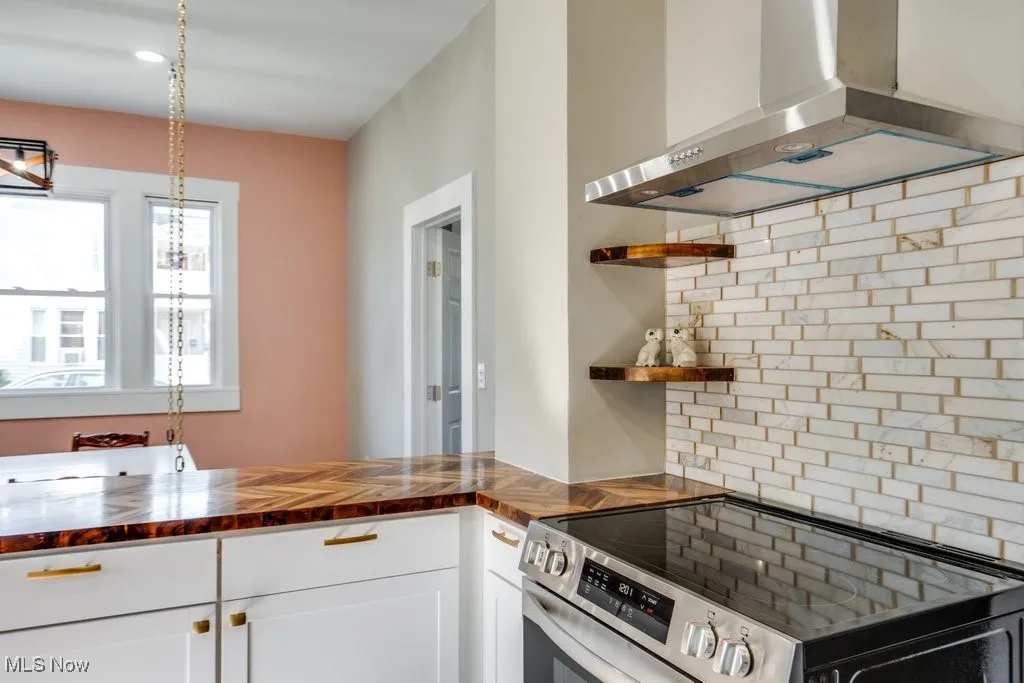 Kitchen featuring wall chimney range hood, stainless steel electric range, white cabinetry, wooden counters, and decorative light fixtures