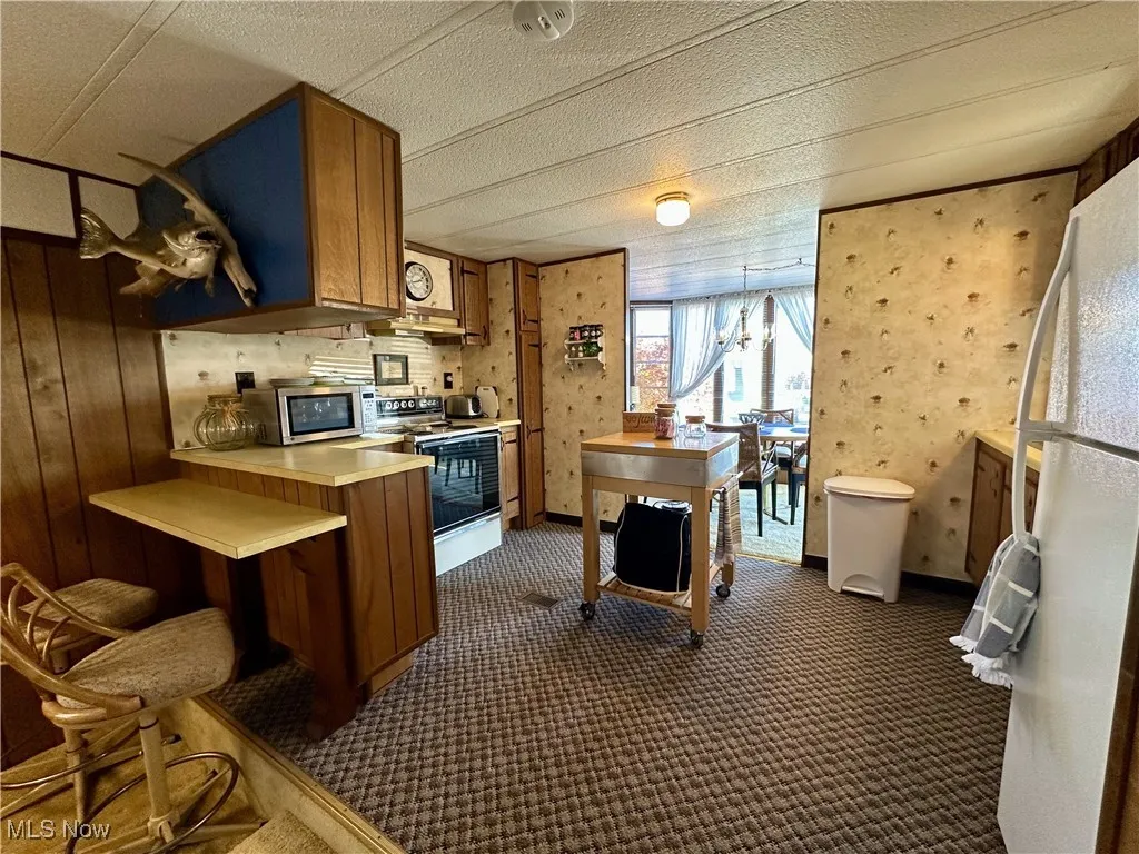 Kitchen featuring dark colored carpet, kitchen peninsula, wooden walls, white appliances, and decorative light fixtures
