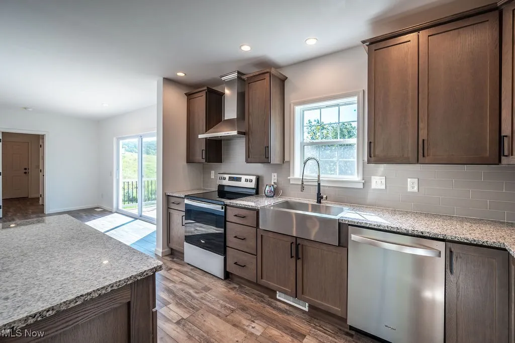 Kitchen with light stone counters, stainless steel appliances, backsplash, wall chimney range hood, and dark wood-type flooring