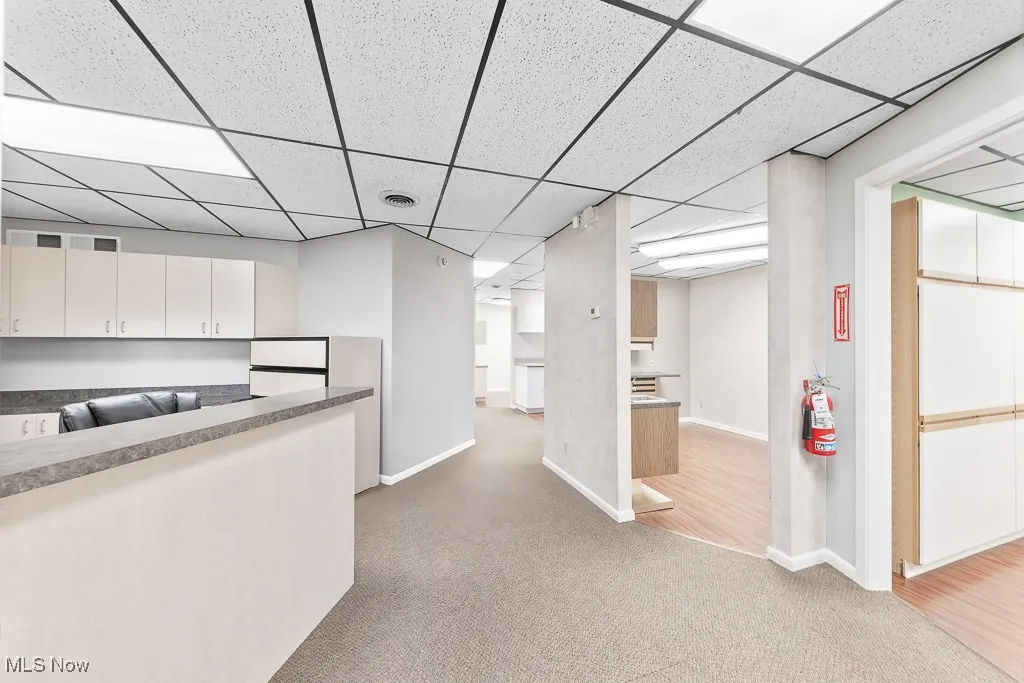 Kitchen with a drop ceiling, white cabinetry, light carpet, and baseboards