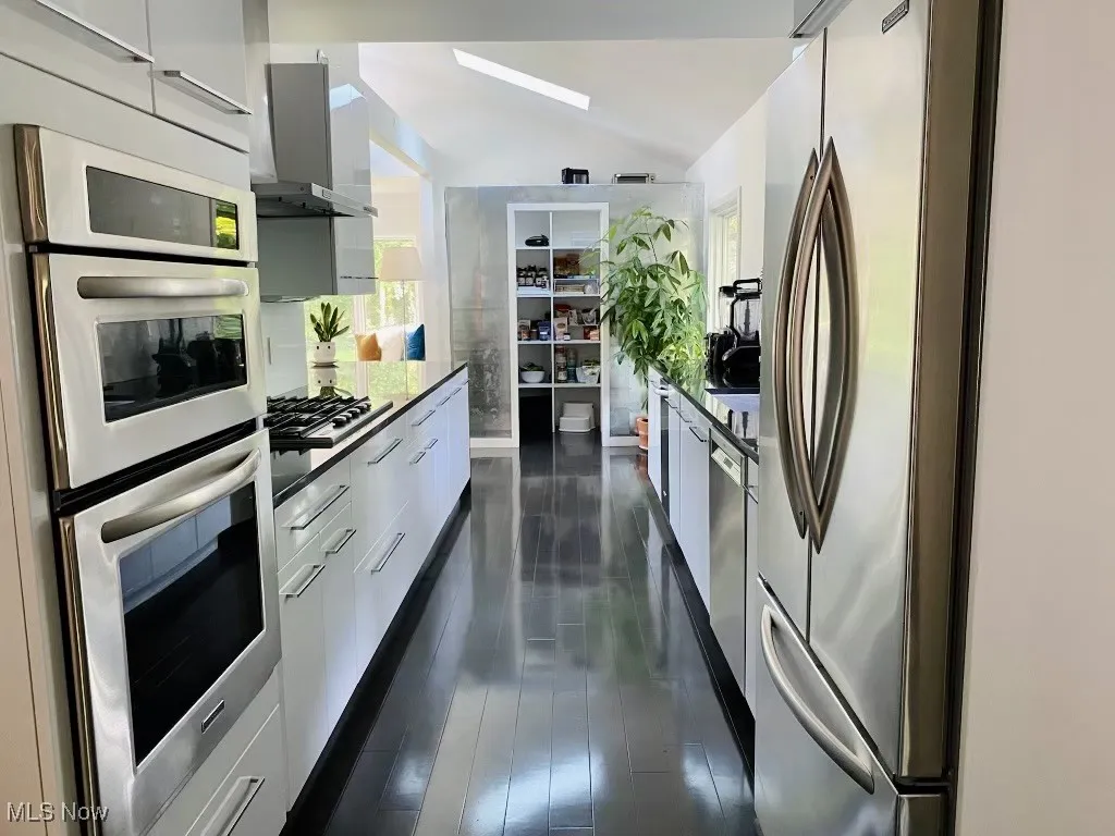 Kitchen featuring lofted ceiling, appliances with stainless steel finishes, wall chimney exhaust hood, and white cabinetry