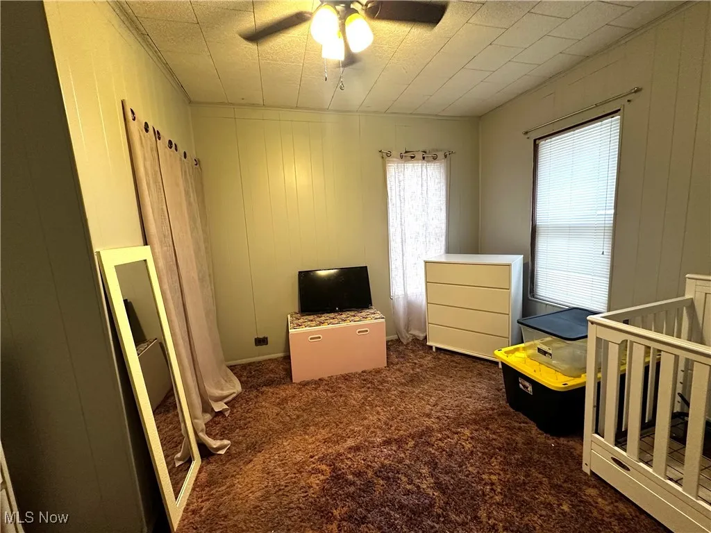 Bedroom with ceiling fan, wooden walls, a crib, and dark colored carpet
