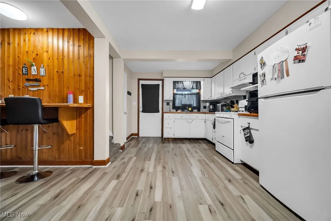 Kitchen with white appliances, light wood-style floors, white cabinets, baseboards, and wooden walls