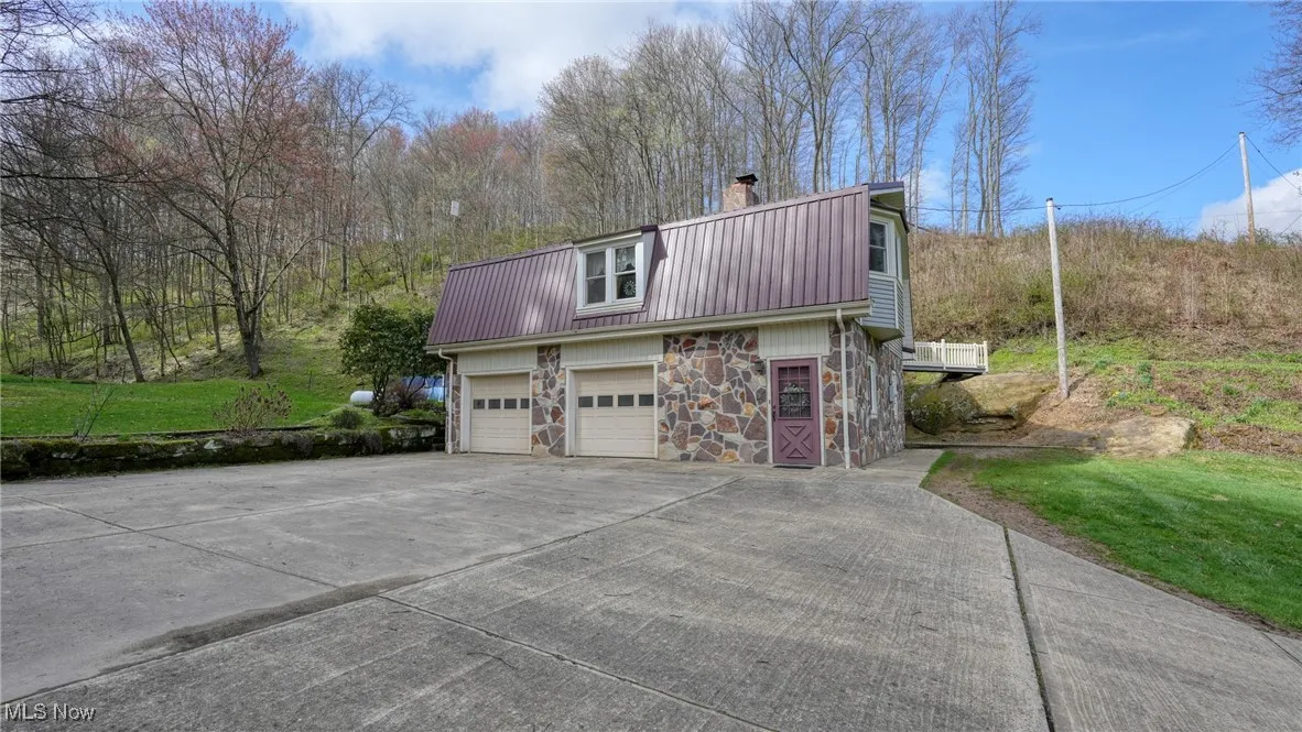 View of side of home with driveway, stone siding, a garage, metal roof, and a gambrel roof