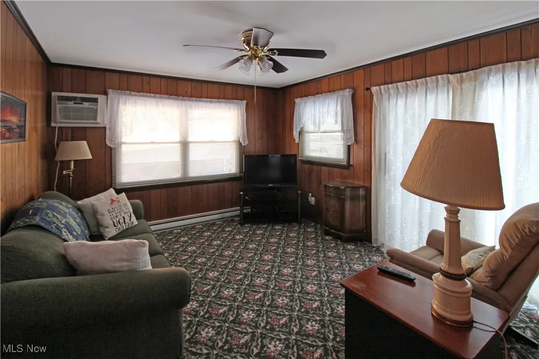 Living room featuring baseboard heating, ceiling fan, wooden walls, and an AC wall unit