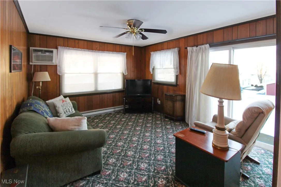 Carpeted living room featuring baseboard heating, plenty of natural light, wooden walls, and an AC wall unit
