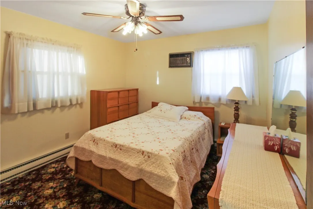 Bedroom featuring multiple windows, a baseboard heating unit, a wall unit AC, and ceiling fan