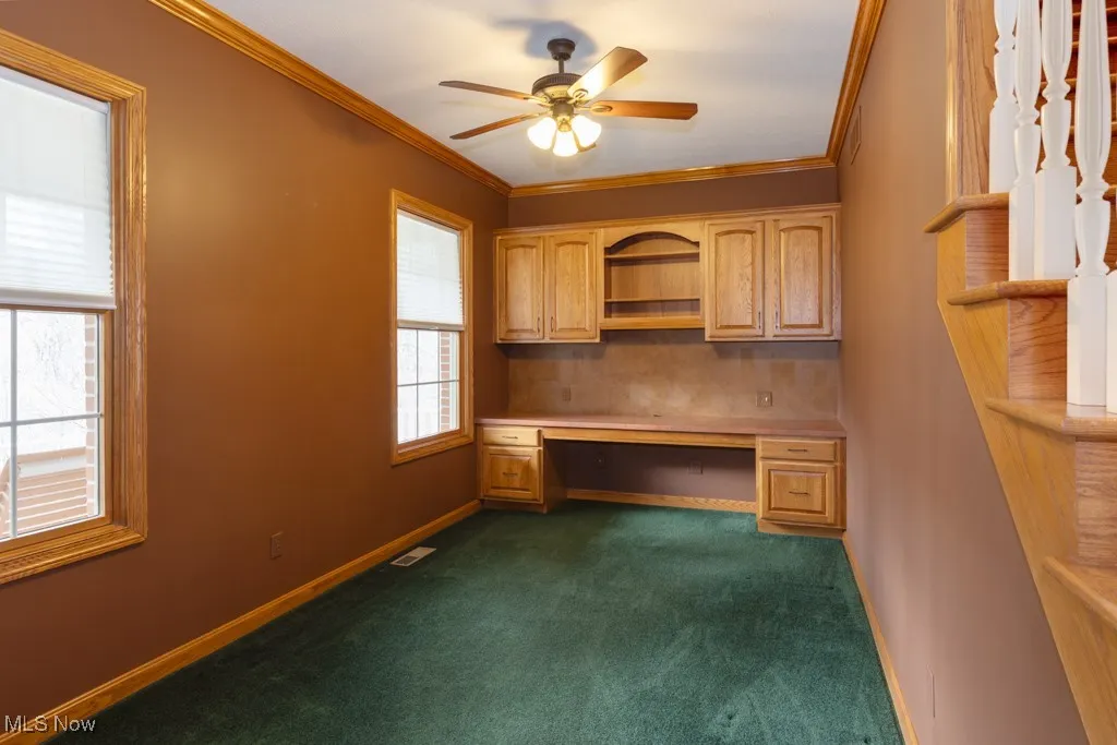 Unfurnished office featuring dark carpet, crown molding, built in desk, and ceiling fan