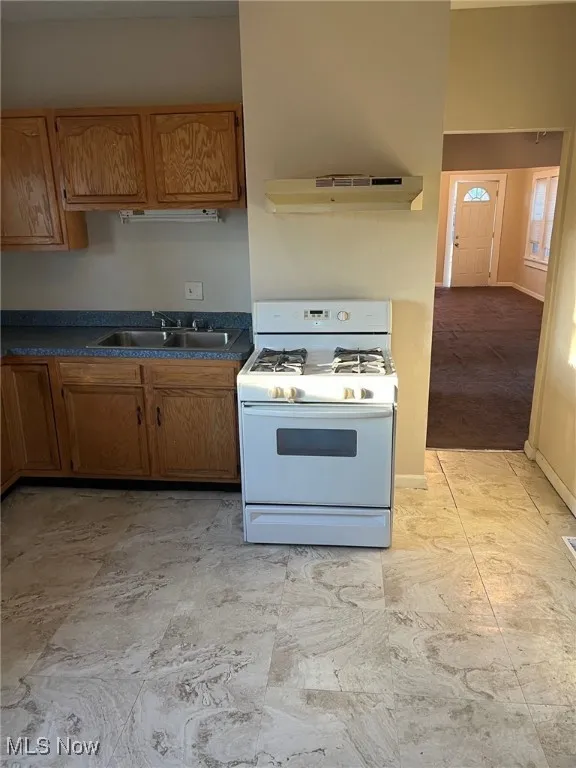 Kitchen featuring brown cabinets, a sink, white gas range, under cabinet range hood, and dark countertops