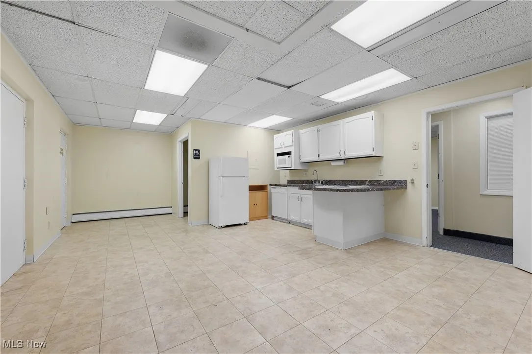 Kitchen featuring white appliances, a baseboard heating unit, dark countertops, a peninsula, and a drop ceiling