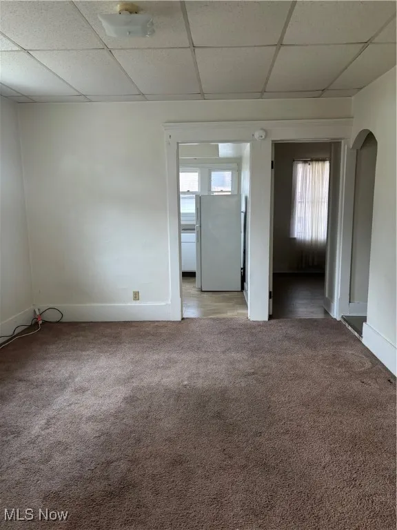 Empty room featuring arched walkways, a drop ceiling, baseboards, and light colored carpet