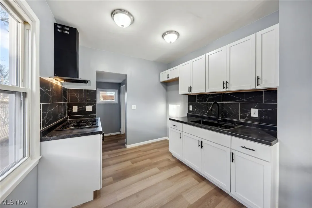 Kitchen with a sink, light wood-type flooring, wall chimney exhaust hood, dark countertops, and cooktop