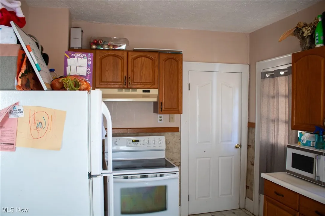 Kitchen featuring a textured ceiling, under cabinet range hood, white appliances, light countertops, and brown cabinetry