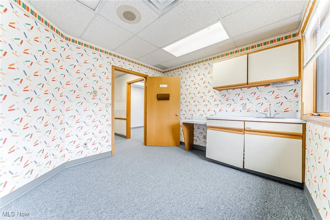 Kitchen with wallpapered walls, carpet, white cabinets, and a drop ceiling