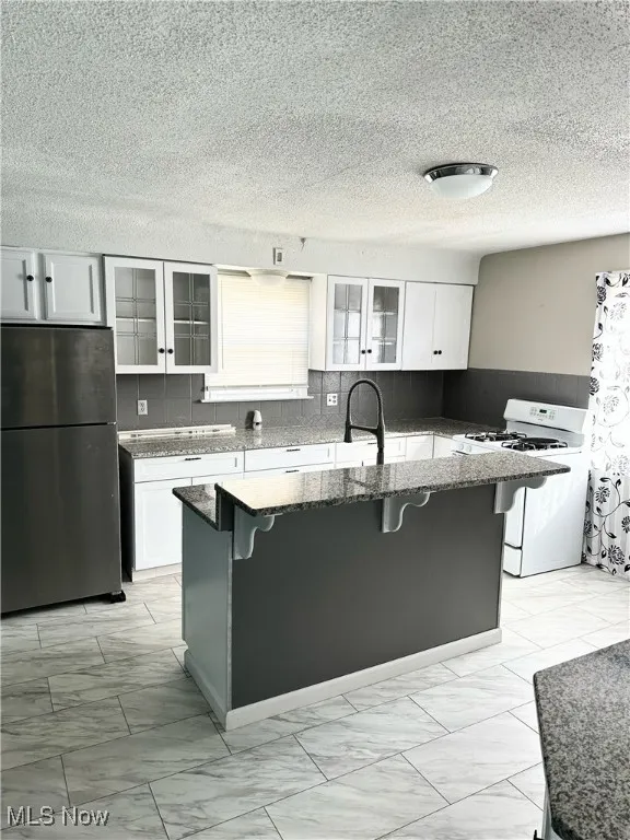 Kitchen with white gas stove, freestanding refrigerator, white cabinets, an island with sink, and a kitchen bar