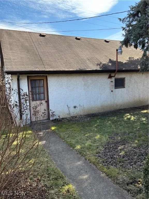 Doorway to property with roof with shingles, a lawn, and stucco siding