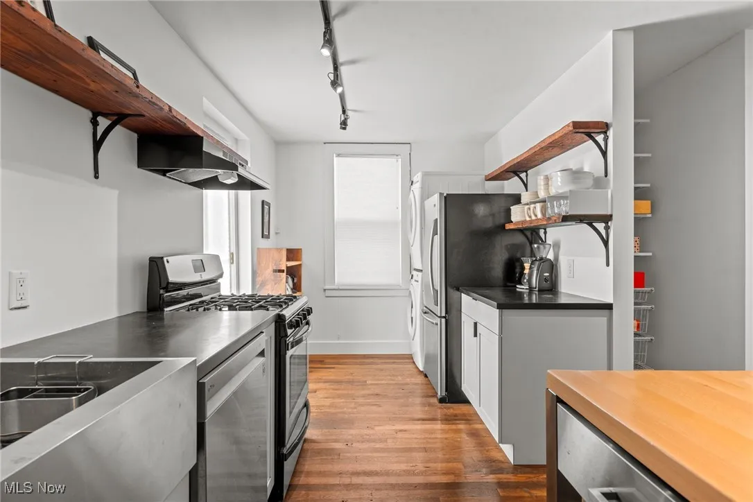 Kitchen with under cabinet range hood, wood finished floors, appliances with stainless steel finishes, open shelves, and dark countertops
