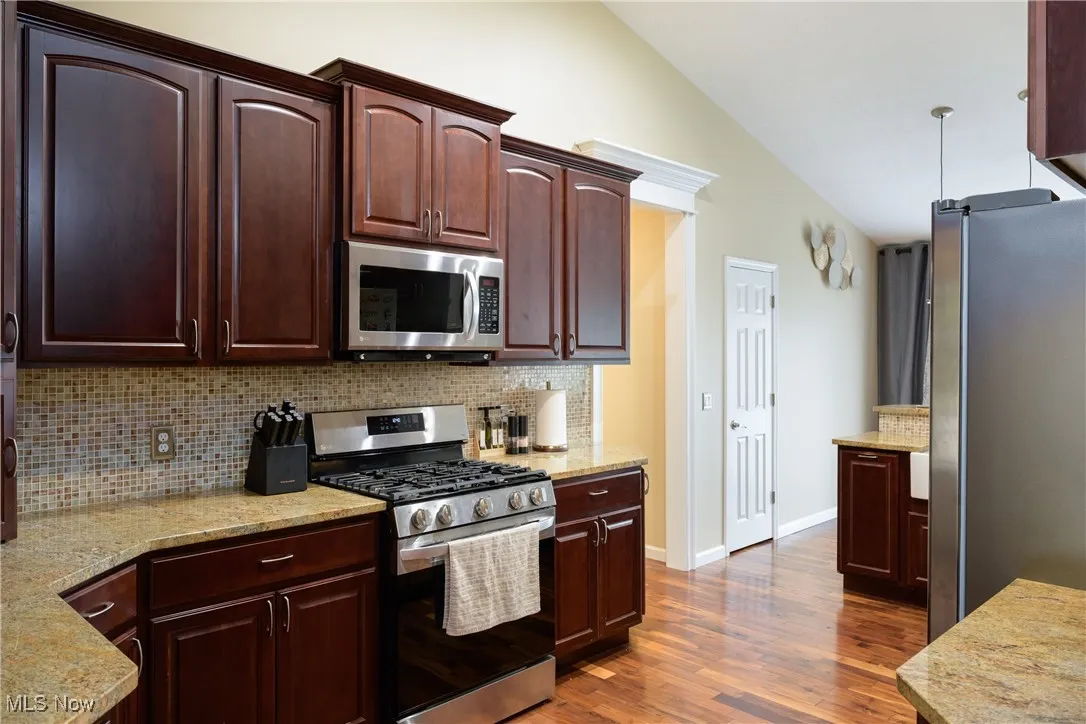 Kitchen featuring tasteful backsplash, light stone countertops, vaulted ceiling, appliances with stainless steel finishes, and dark wood-style floors