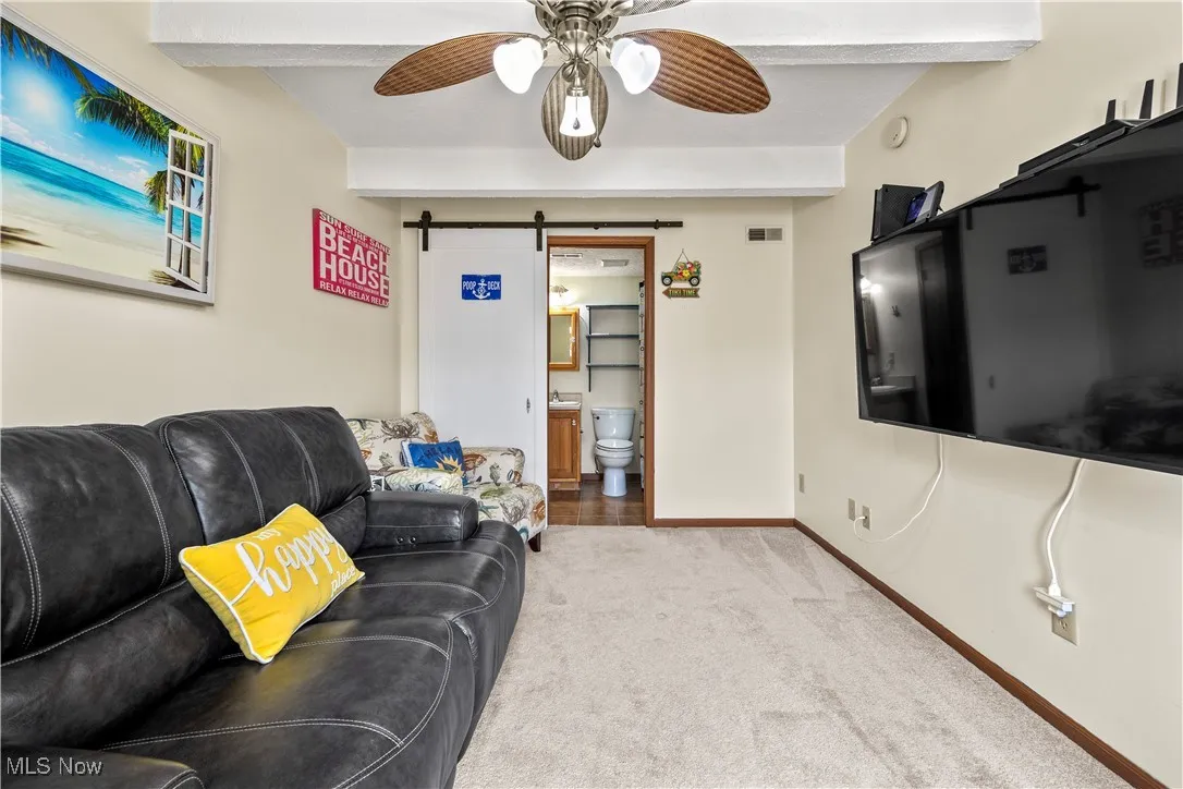Carpeted living area with visible vents, baseboards, beam ceiling, ceiling fan, and a barn door