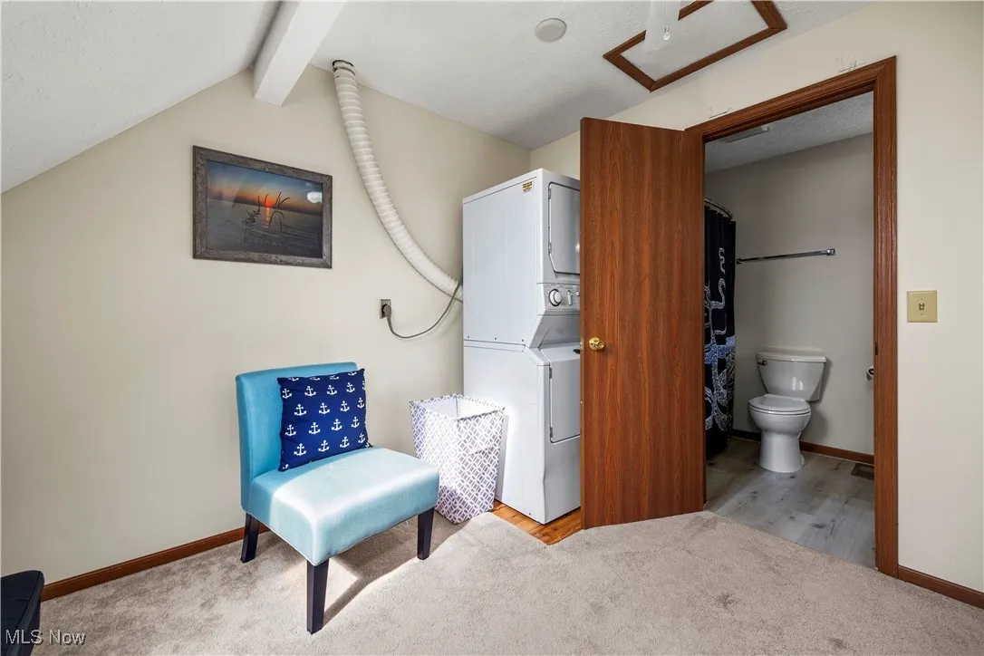 Living area featuring vaulted ceiling with beams, baseboards, carpet, and stacked washer and dryer