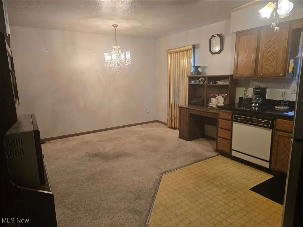 Kitchen with baseboards, light carpet, dishwasher, dark countertops, and a notable chandelier