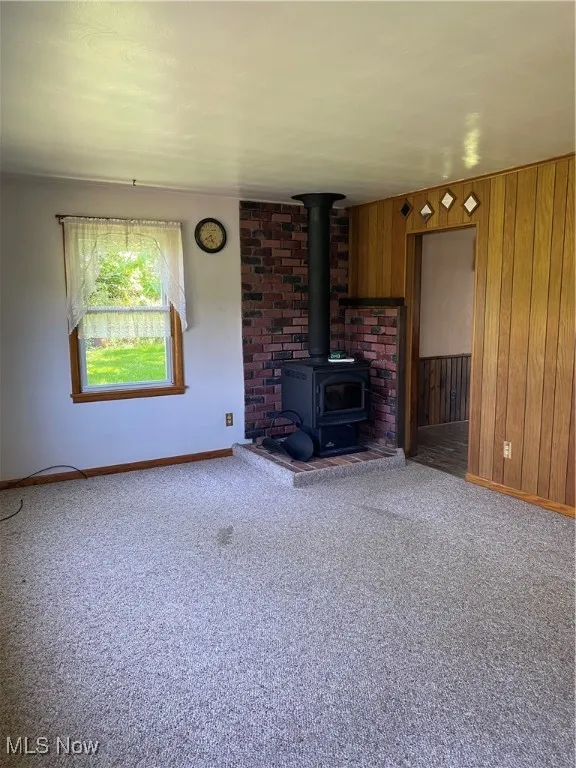 Unfurnished living room featuring baseboards, carpet, a wood stove, and wooden walls