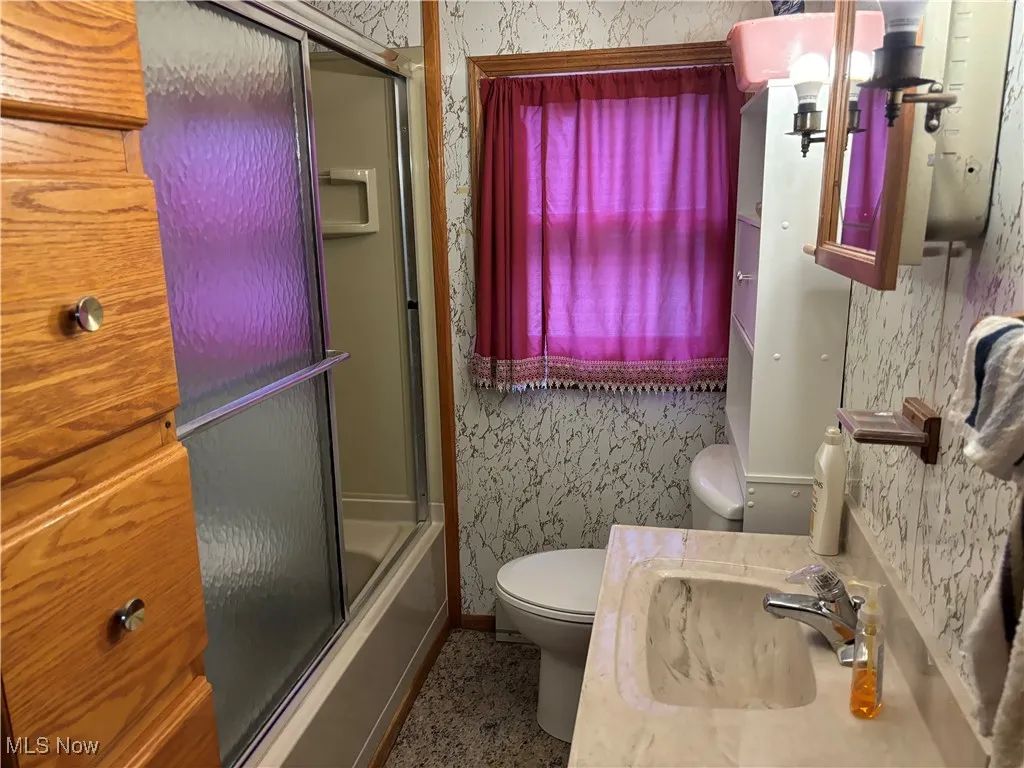 Bathroom with wallpapered walls, baseboards, toilet, combined bath / shower with glass door, and vanity