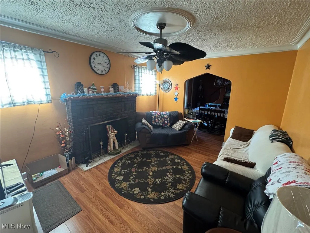 Living area with a textured ceiling, crown molding, a brick fireplace, wood finished floors, and a ceiling fan